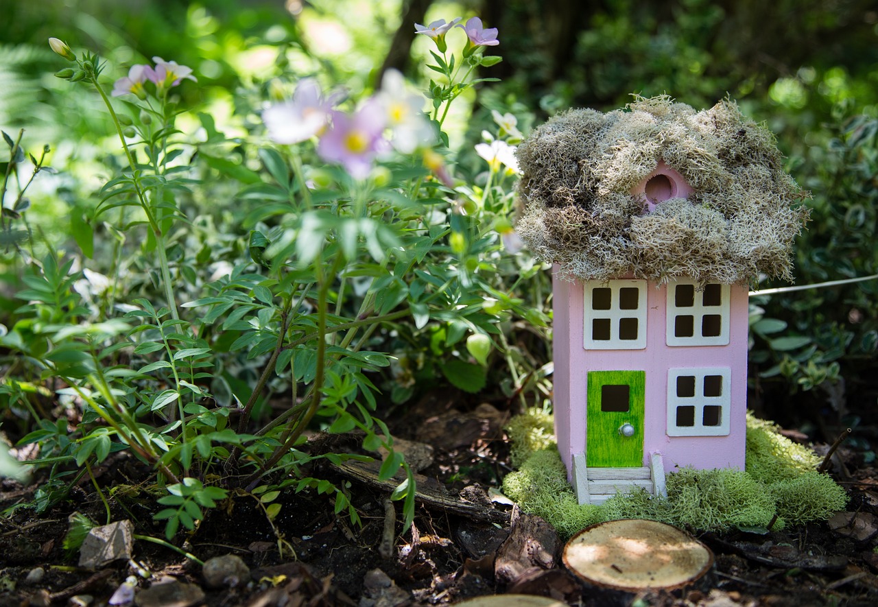 A wooden fairy house in a garden next to a flowering plant.