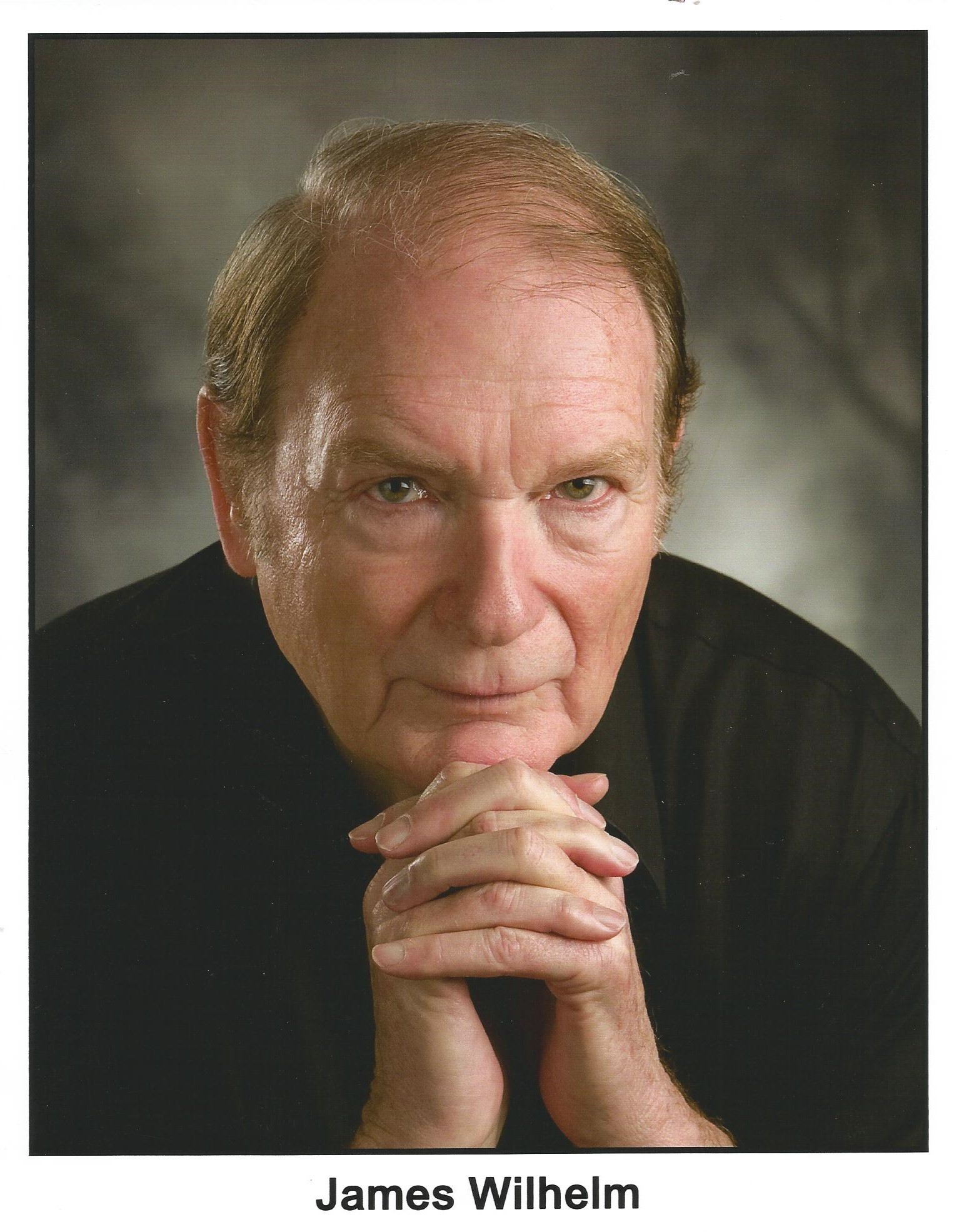 A man in a dark turtleneck shirt.  He is leaning forward with his chin resting on his folded hands.