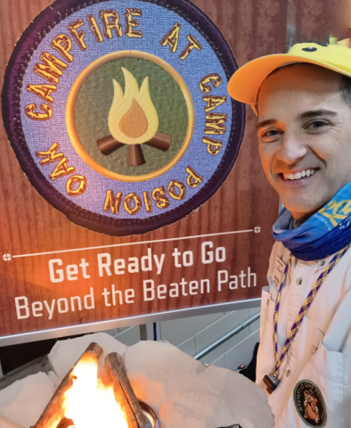 A man wearing a yellow ballcap and dressed as a summer camp counselor, wearing a whistle around his neck, standing next to a banner displaying an image of a Camp Poison Oak merit badge and the words Get Ready to Go Beyond the Beaten Path