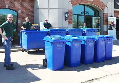Three men in green shirts standing next to a bank of blue plastic recycling toters.