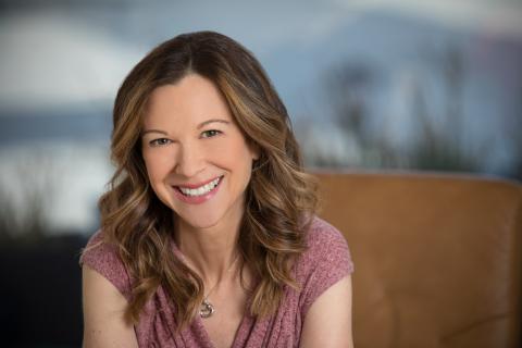 A woman with long, wavy brown hair is wearing a pink shirt and smiling.