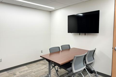 A small meeting room with one table and four chairs.  A computer monitor is mounted on the wall.