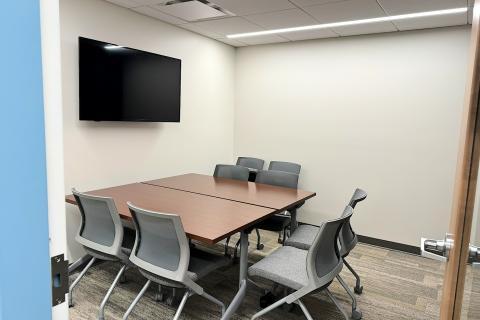A small meeting room with two tables and eight chairs.  A computer monitor/TV is mounted on the wall.