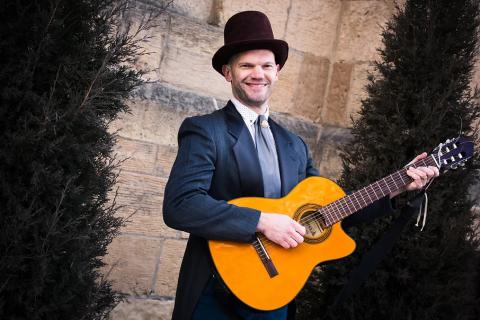 A man wearing a top hat and formal coat and tie is posing with guitar in front of a stone wall.