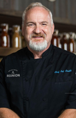 A man with grey hair, a mustache, and beard is wearing a black chef's uniform with "Reunion" and "Chef Art Smith" embroidered on the jacket.