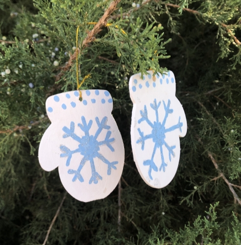 Wooden ornaments in the shape of mittens painted with a blue snowflake and dots on a white background hanging in a tree.