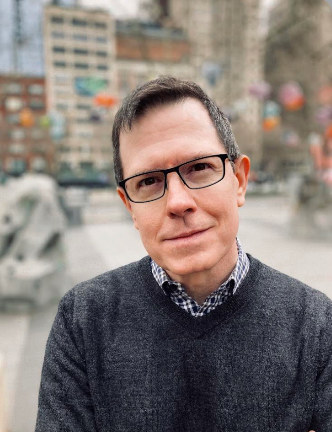 A man with short hair and glasses wearing a dark sweater, with a blurry cityscape in the background.