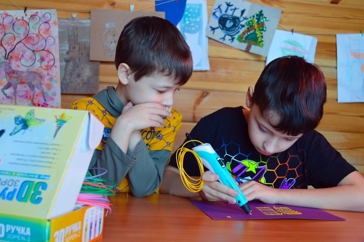 One elementary-aged boy is creating something using a hand-held 3D printing pen while another boy watches intently.