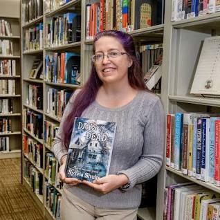 A woman with glasses and long purple hair, holding a book, and standing next to a library bookshelf.