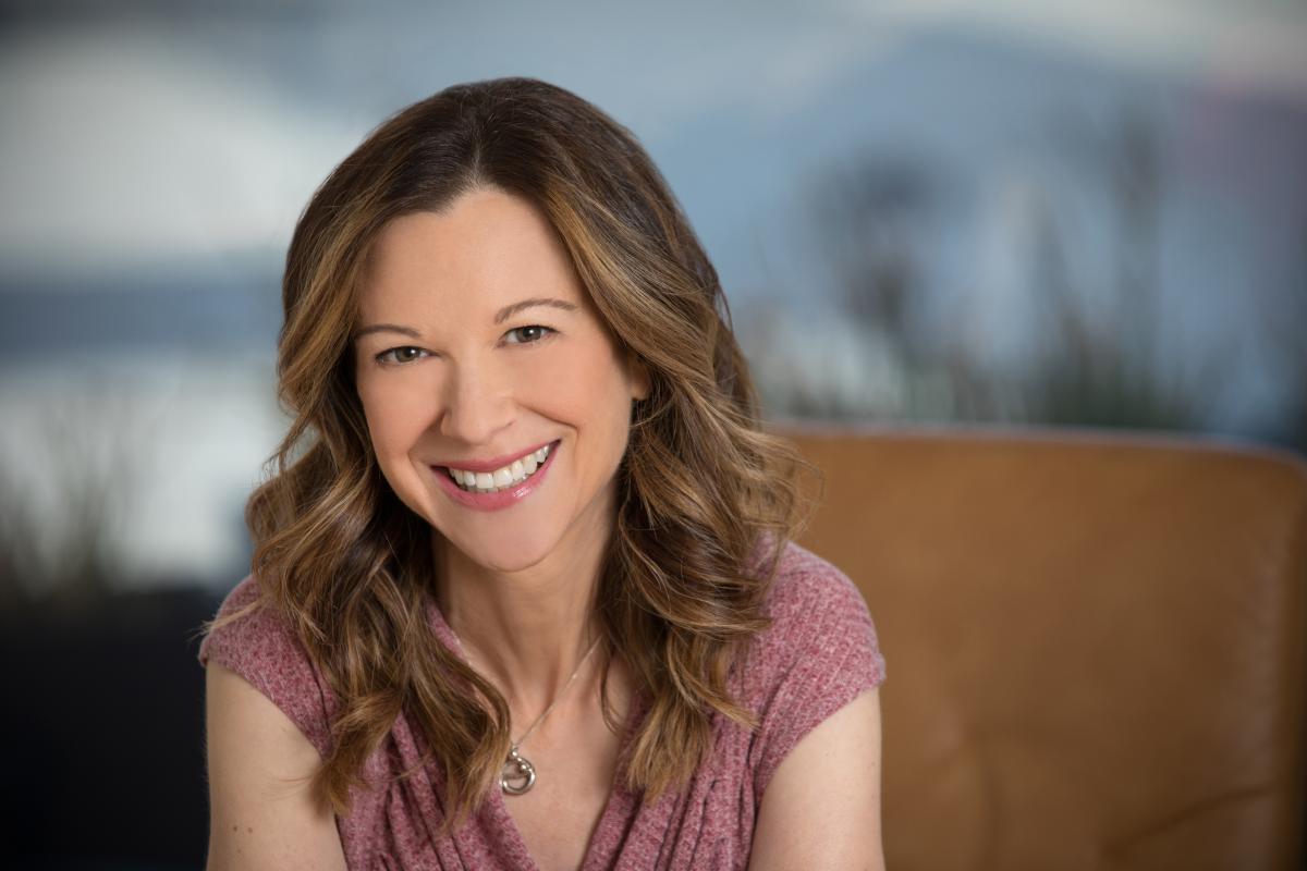 A woman with long, wavy brown hair is wearing a pink shirt and smiling.