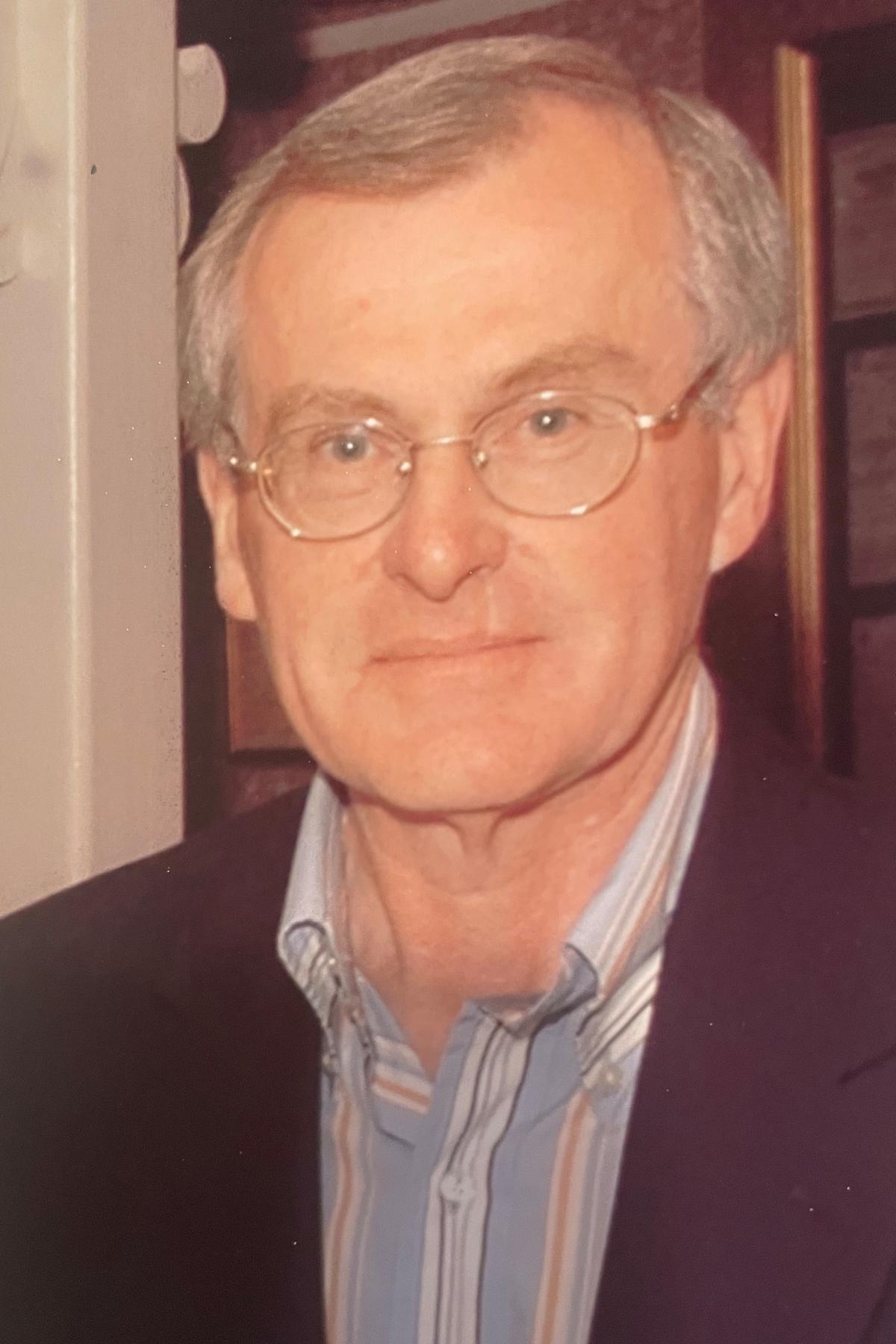 A man with graying hair and glasses is wearing a striped button-down shirt and a dark blazer.  The collar of his shirt is open.  He is not wearing a tie.