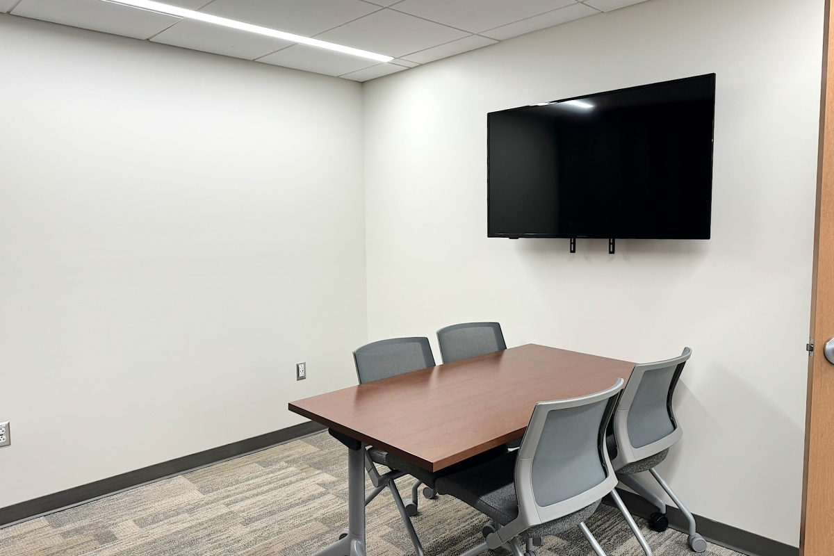 A small meeting room with one table and four chairs.  A computer monitor is mounted on the wall.