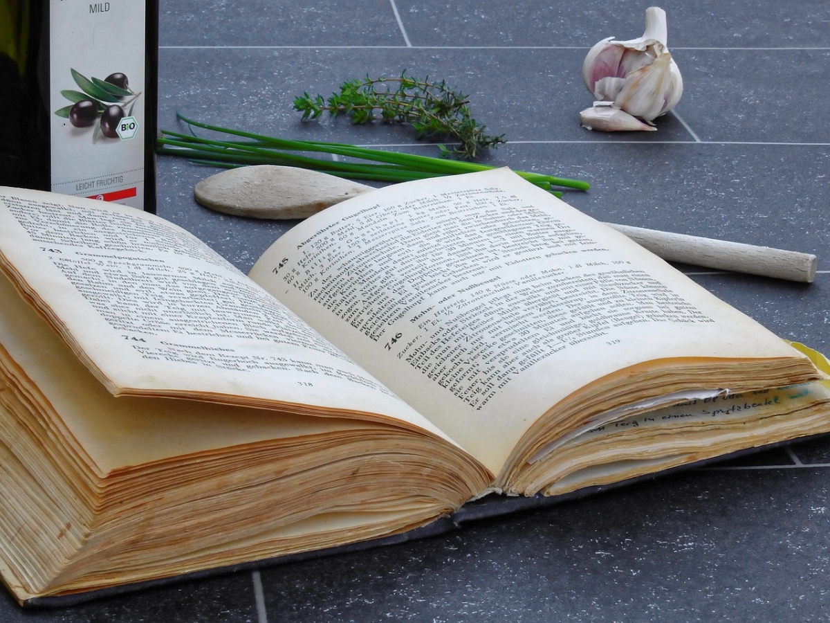 An open cookbook on a countertop next to a wooden spoon, a clove of garlic, and some parsley.